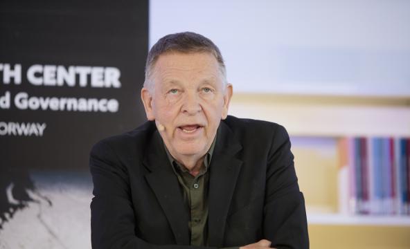 Petter Nore lener seg på et podie mens han tydeligvis snakker til et publikum. Vi ser bøker og en flier med High North Center bak ham.