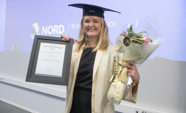 Female student, wearing a graduation hat, holding a diploma and flowers while smiling into the camera.