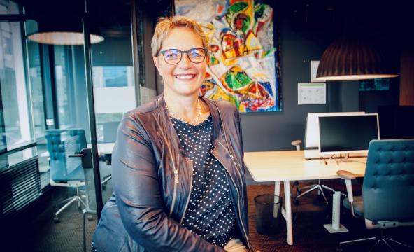 Kvinne ser i kamera og smiler. I bakgrunnen er det et fargerikt kunstverk. 