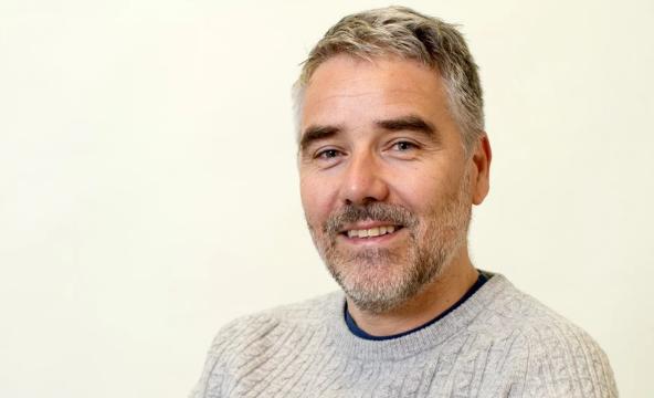 Portrettbilde av mann i genser med grått hår og tredagers skjegg. Foto