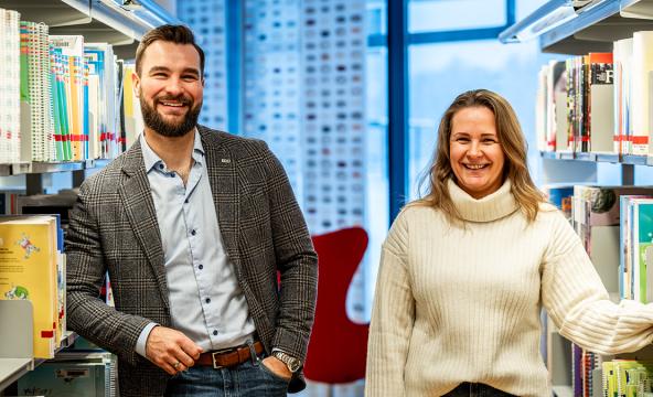 To smilende studenter står mellom bibliotekhyller