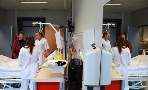 Sykepleierstudenter i uniform sammen med rektor ved en sykeseng i et øvingsrom. Foto