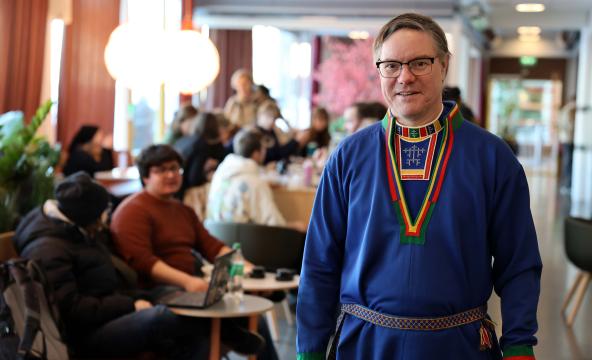 Voksen mann i samekofte i en kantine med mange mennesker i bakgrunn. Foto