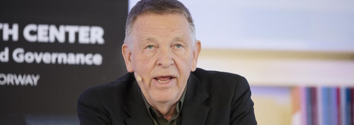 Petter Nore lener seg på et podie mens han tydeligvis snakker til et publikum. Vi ser bøker og en flier med High North Center bak ham.