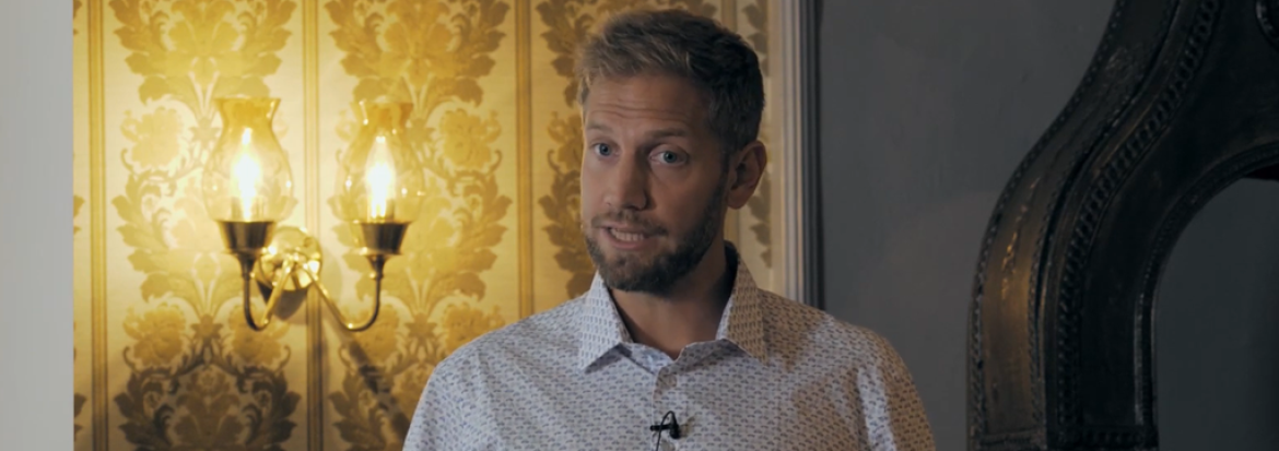 Andreas Østhagen holder foredrag foran en vegg med gult teppe som har en lampett på seg. 