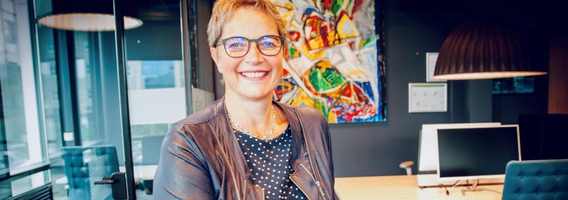 Kvinne ser i kamera og smiler. I bakgrunnen er det et fargerikt kunstverk. 