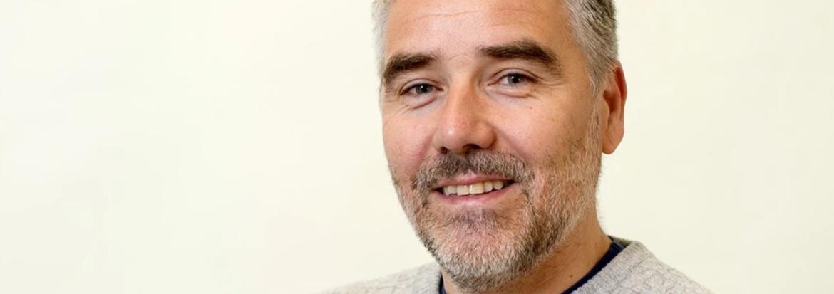 Portrettbilde av mann i genser med grått hår og tredagers skjegg. Foto