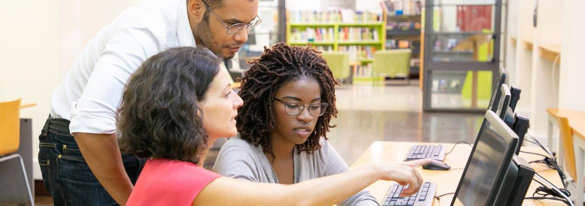 Three students, one male and to female, in front of a computer at a library, one is pointing at the screen.