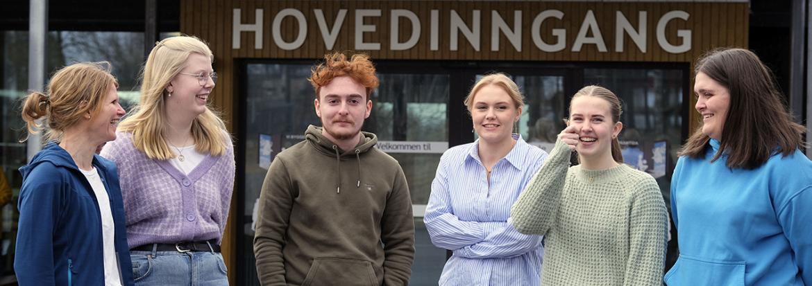 Seks lærerstudenter og ansatte på rekke foran inngangen på et bygg. Grunnskolelærer, grunnskolelærerutdanning. Foto