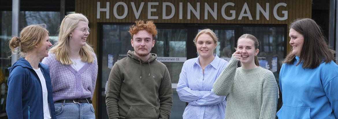 Seks mennesker på rekke og rad foran en hovedinngang. Foto