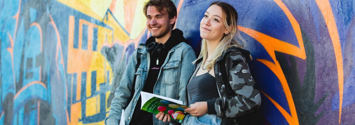 To studenter står og lener seg mot en vegg med grafitti. Foto.