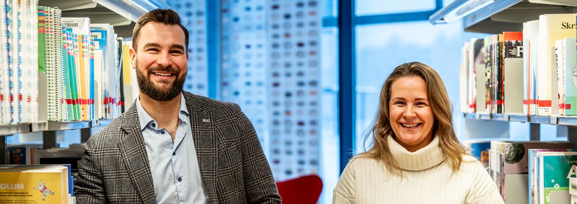 To smilende studenter står mellom bibliotekhyller