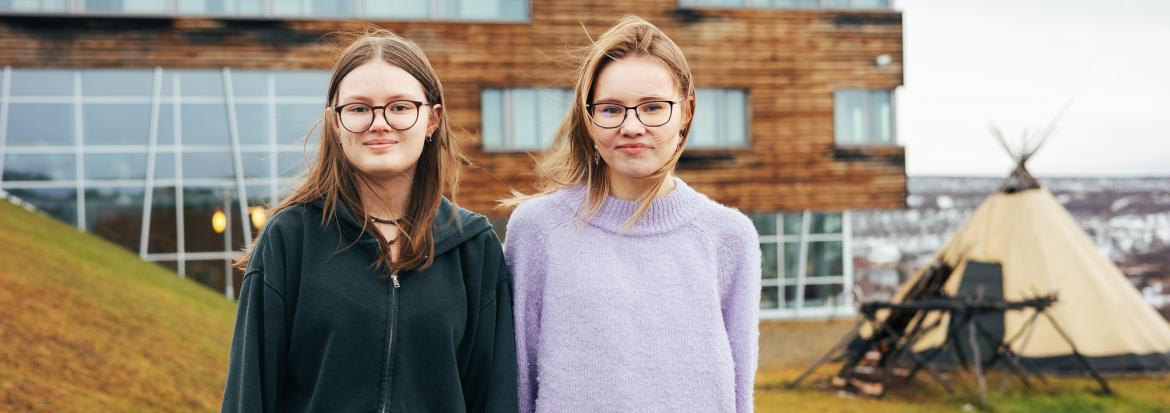 To unge jenter står foran en bygning og ser inn i kamera.