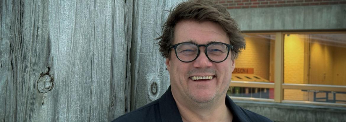 Voksen mann med mørkt hår og briller hvilende ved en vegg med universitetsbygg i bakgrunnen. Halvard Benjaminsen