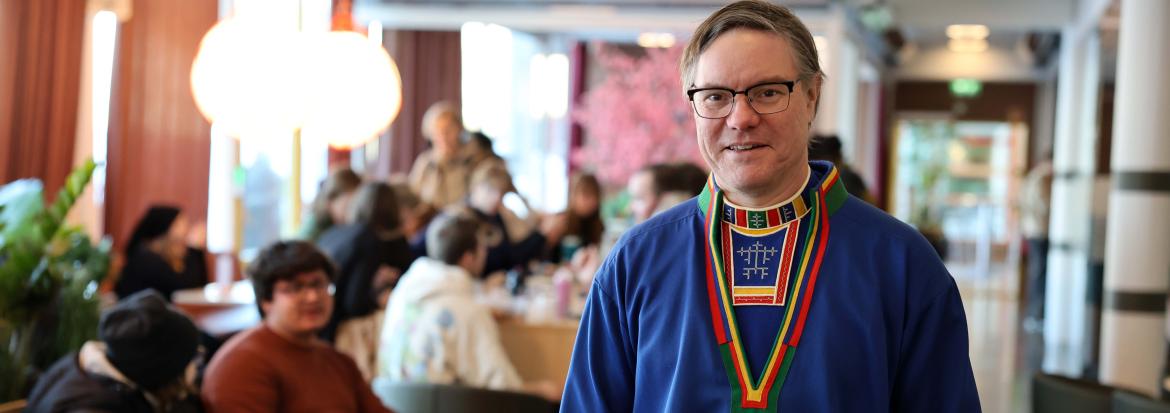 Voksen mann i samekofte i en kantine med mange mennesker i bakgrunn. Foto