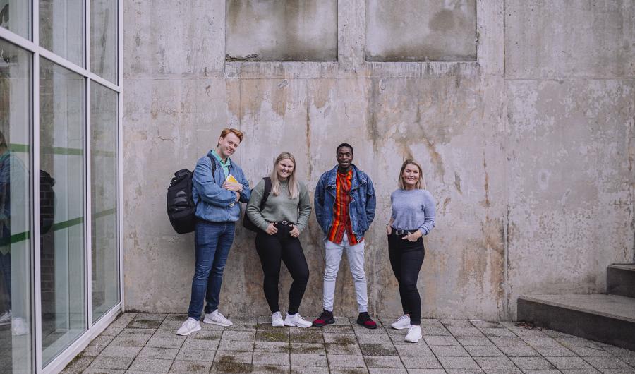 Fire studenter står ved en grå vegg og ser inn i kamera