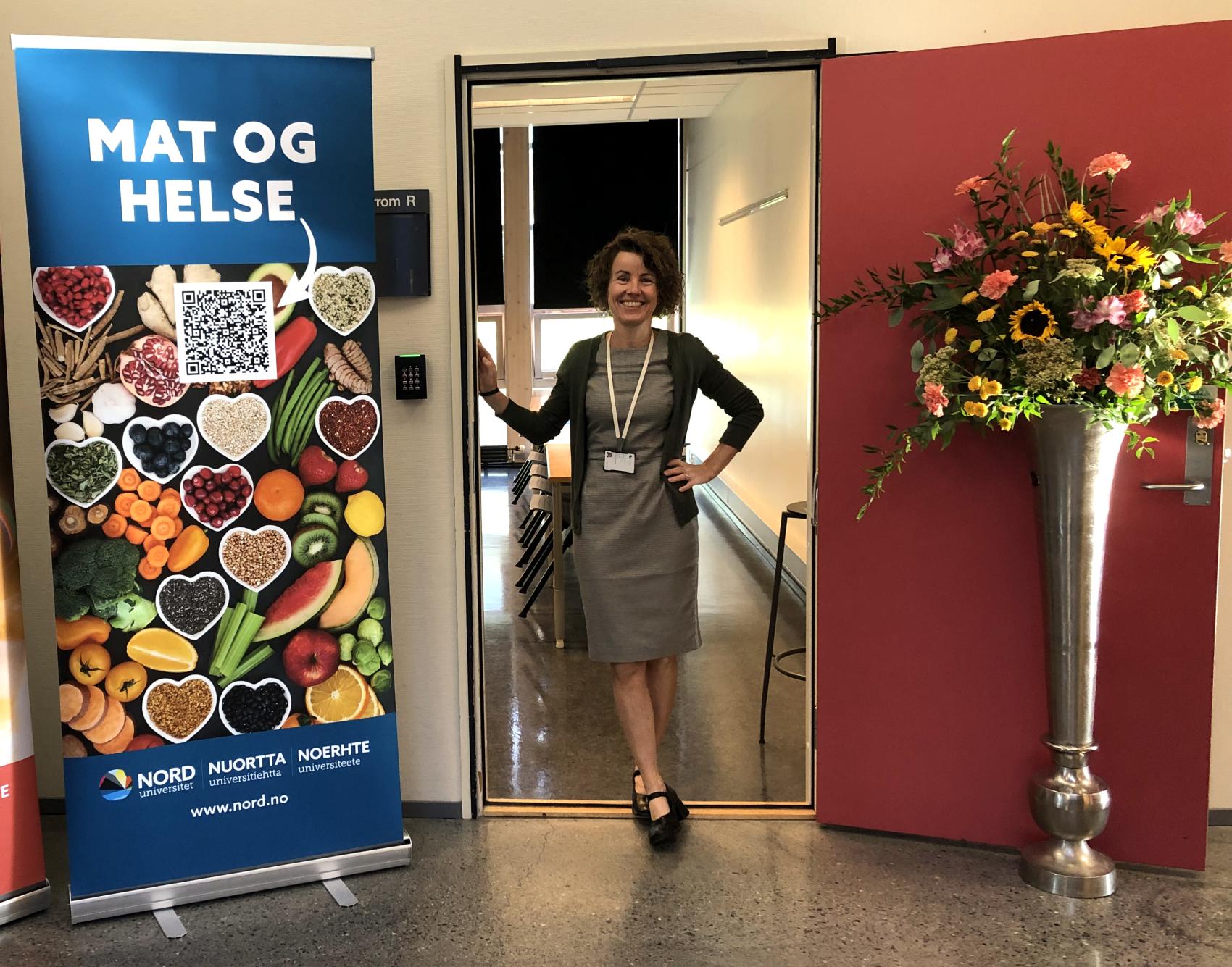 Camilla Sandvik avbildet i en døråpning, flankert av plakat for studiet Mat og helse og blomster