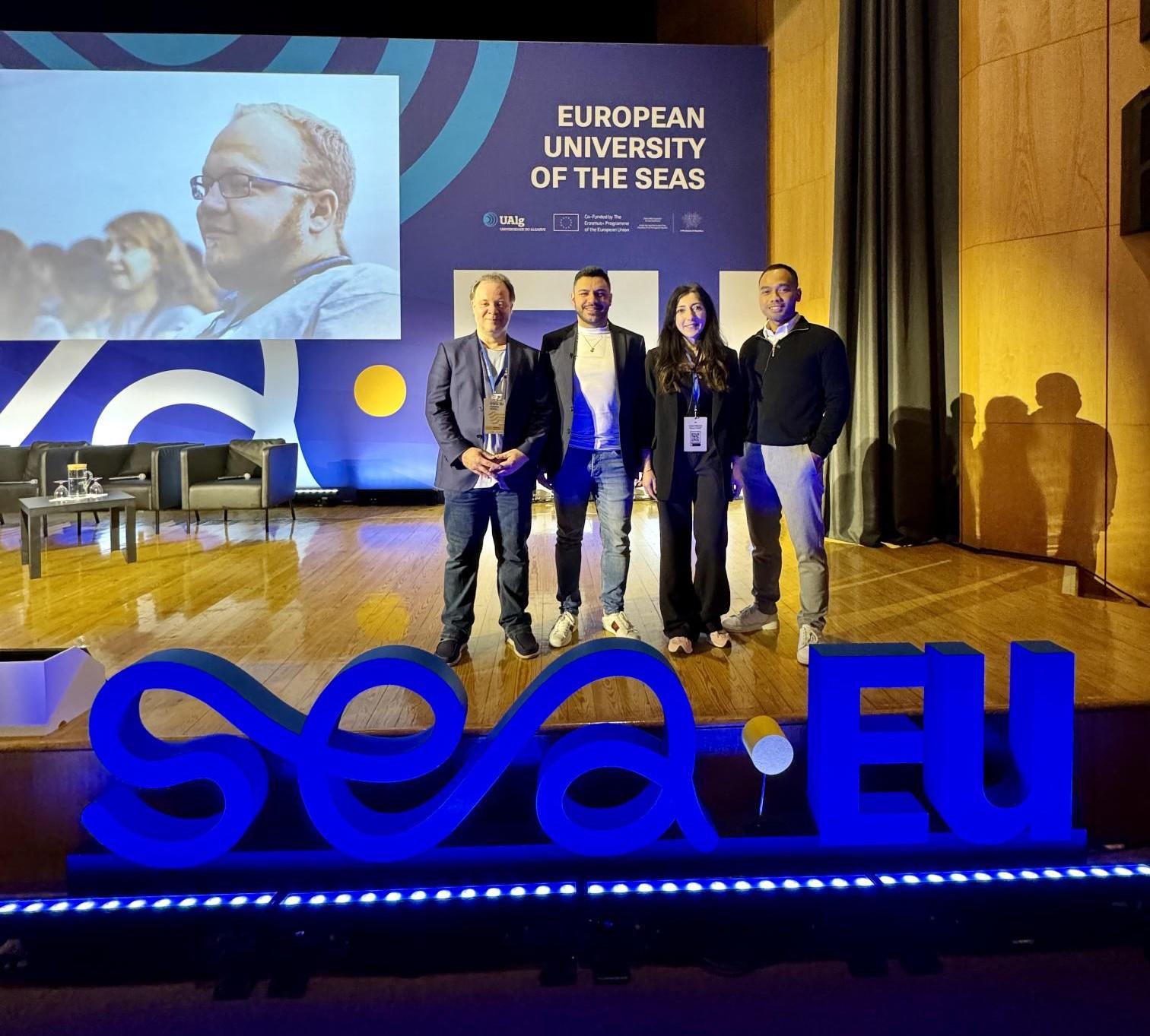 Fire personer står på en scene, foran et blått lysende SEA-EU-skilt