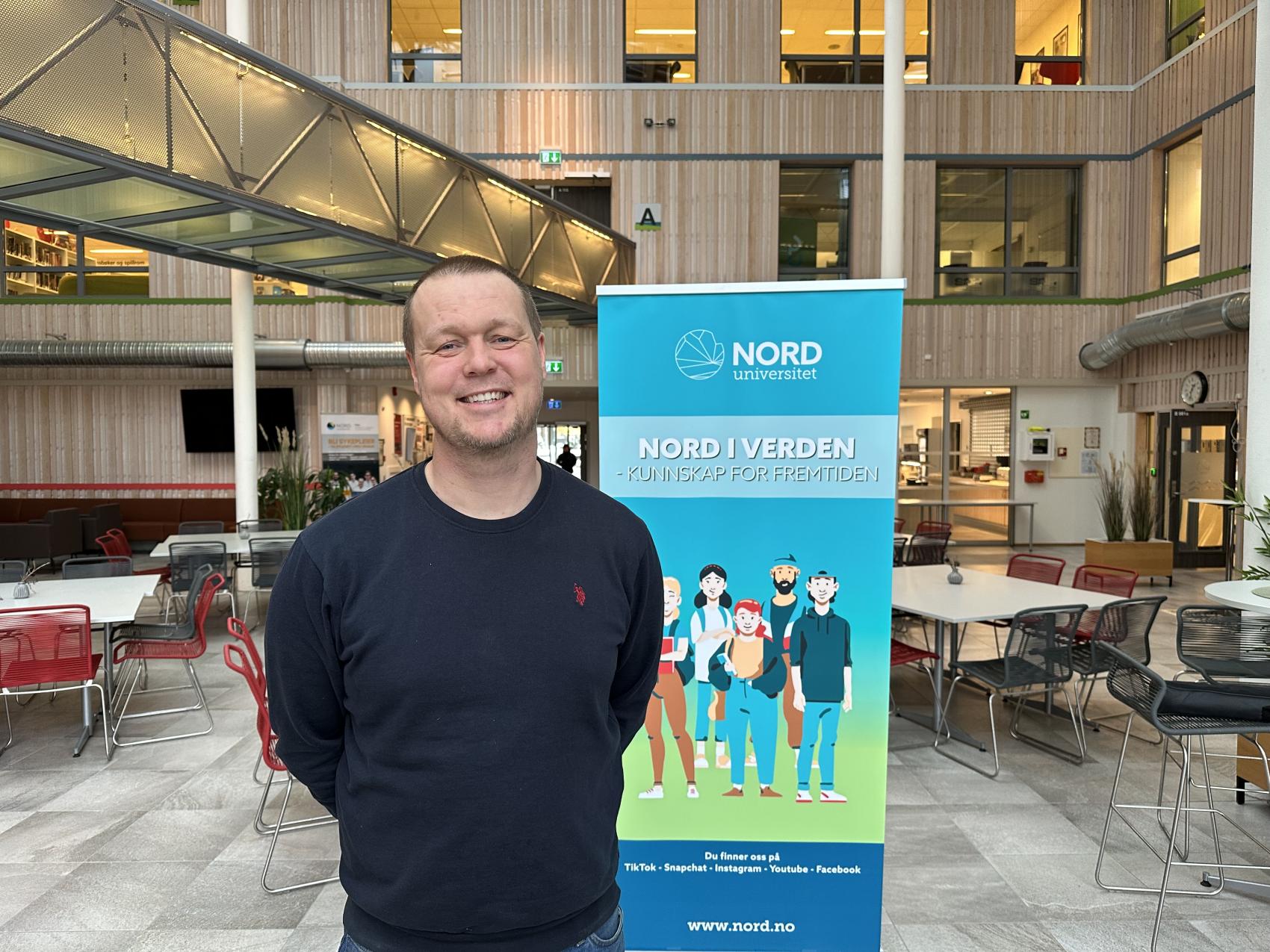 Mann står innendørs foran en reklameplakat. Foto.