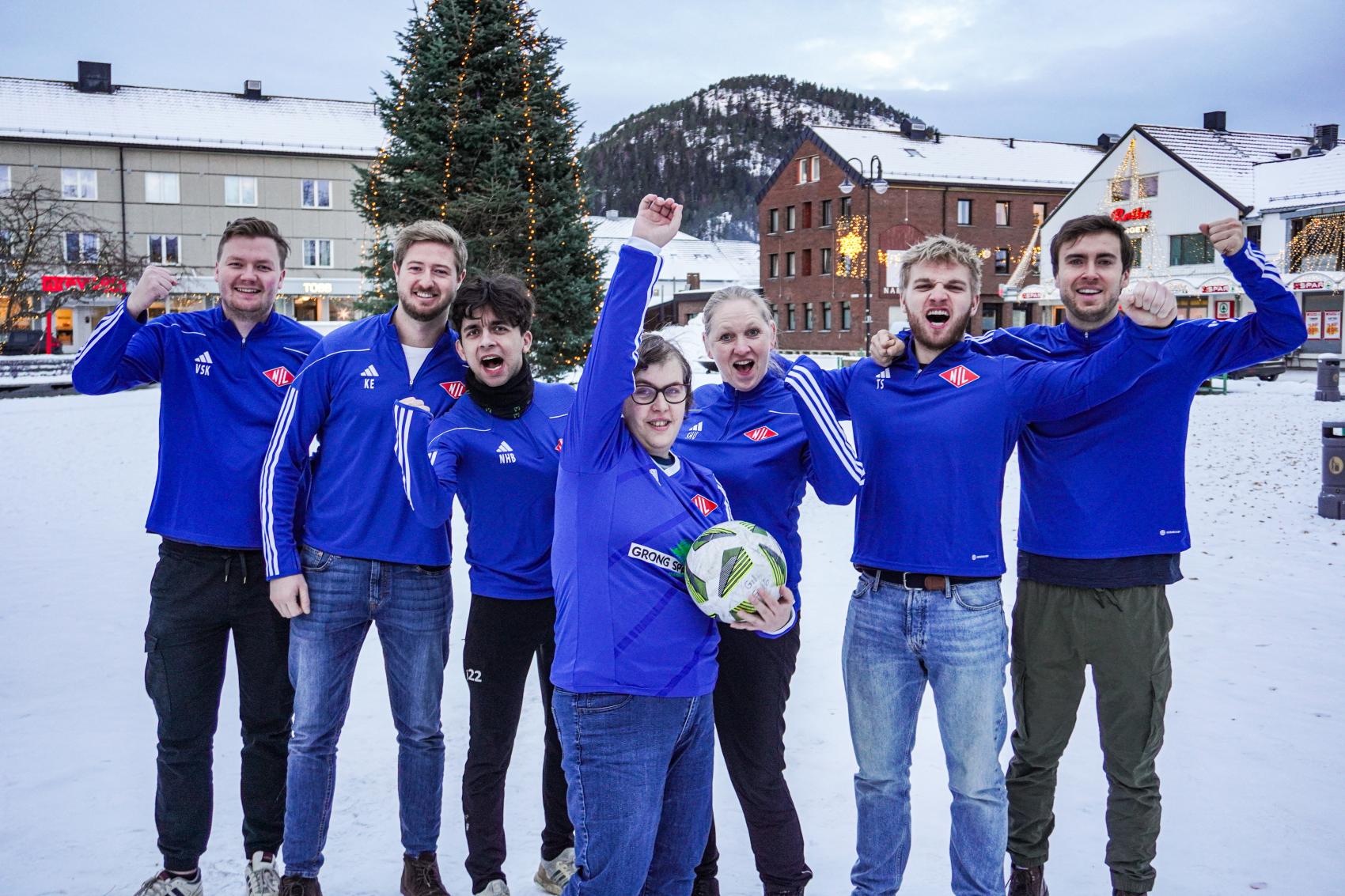 Fotballspillere i blå trøyer som jubler med hendene over hodet. Alle står i sentrum av en by på vinteren. Foto