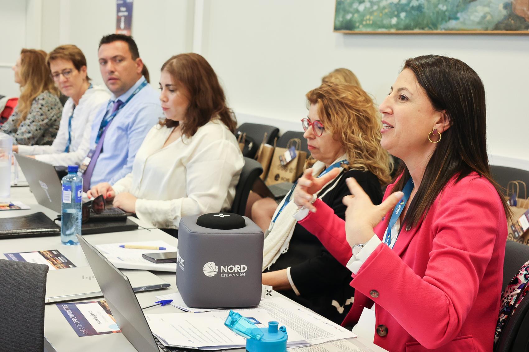A woman dressed in pink at the front, talking actively in a meeting