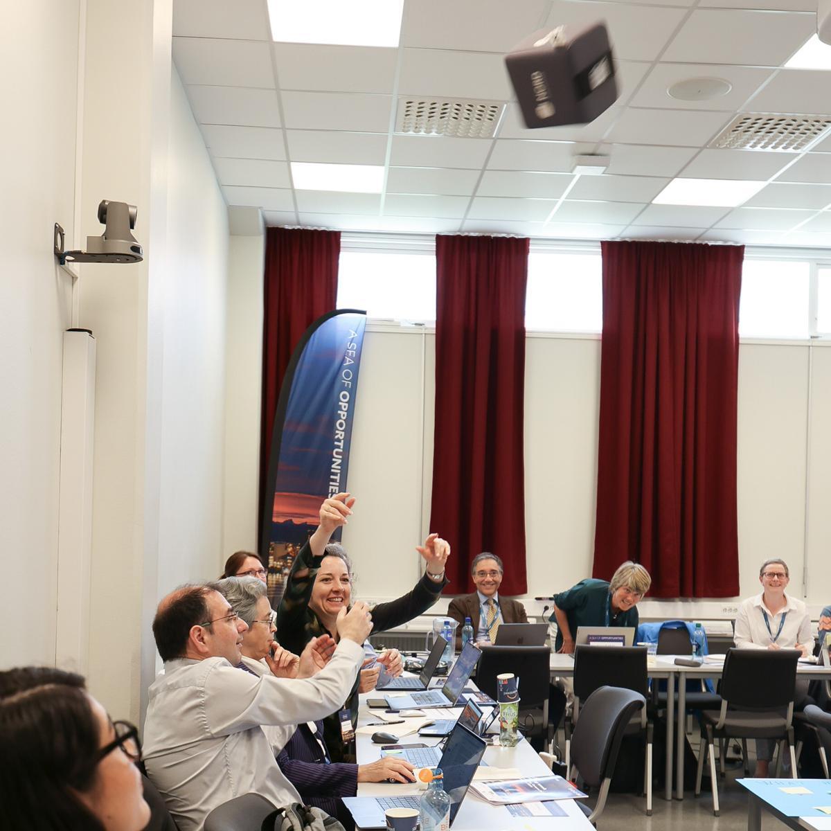 People in a meeting, sitting behind a row of desks. A woman is throwing a microphone cube in the air, the other participants looking at it.