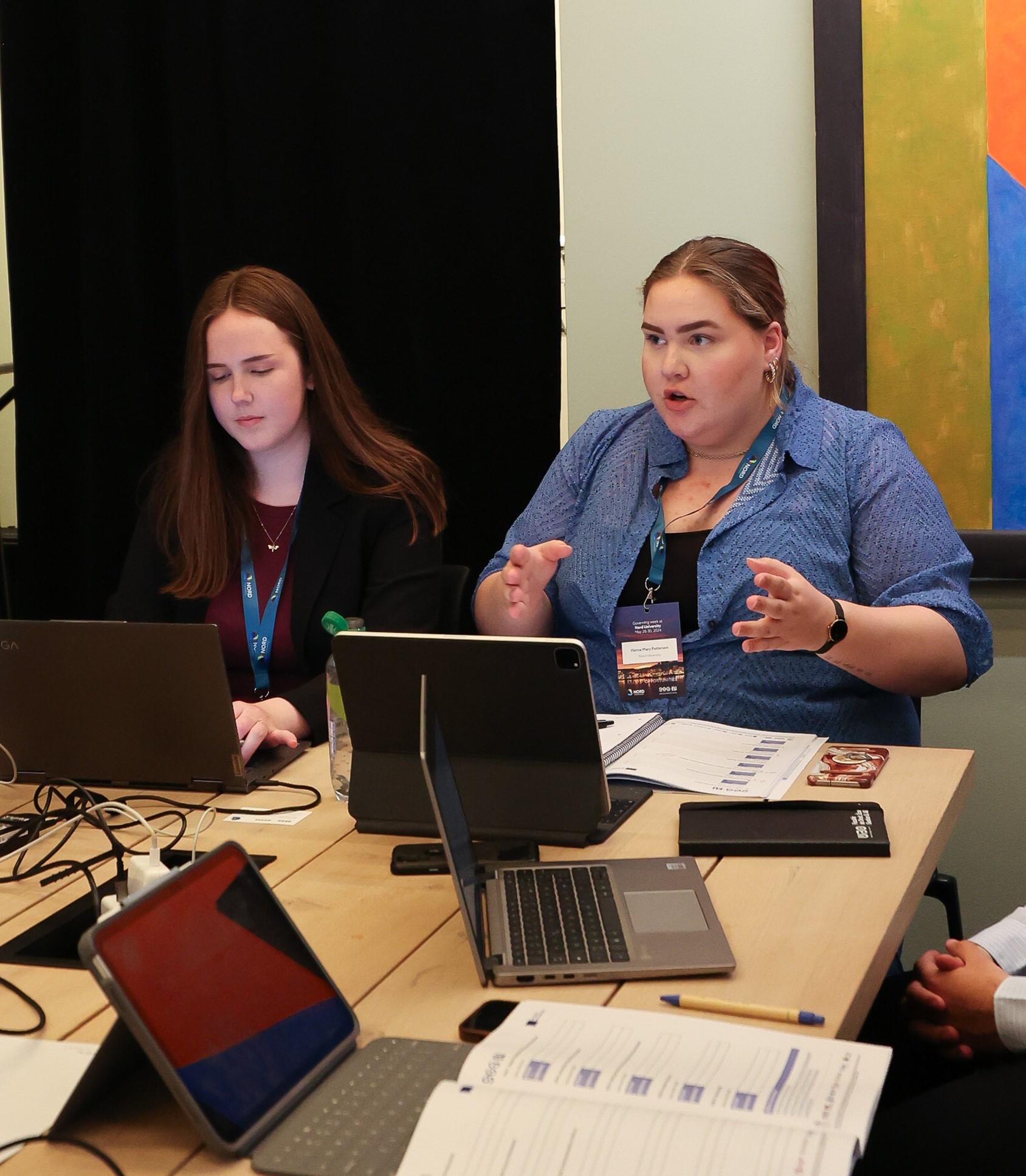 Two young women sitting by a table with laptops in front of them. One of them is talking