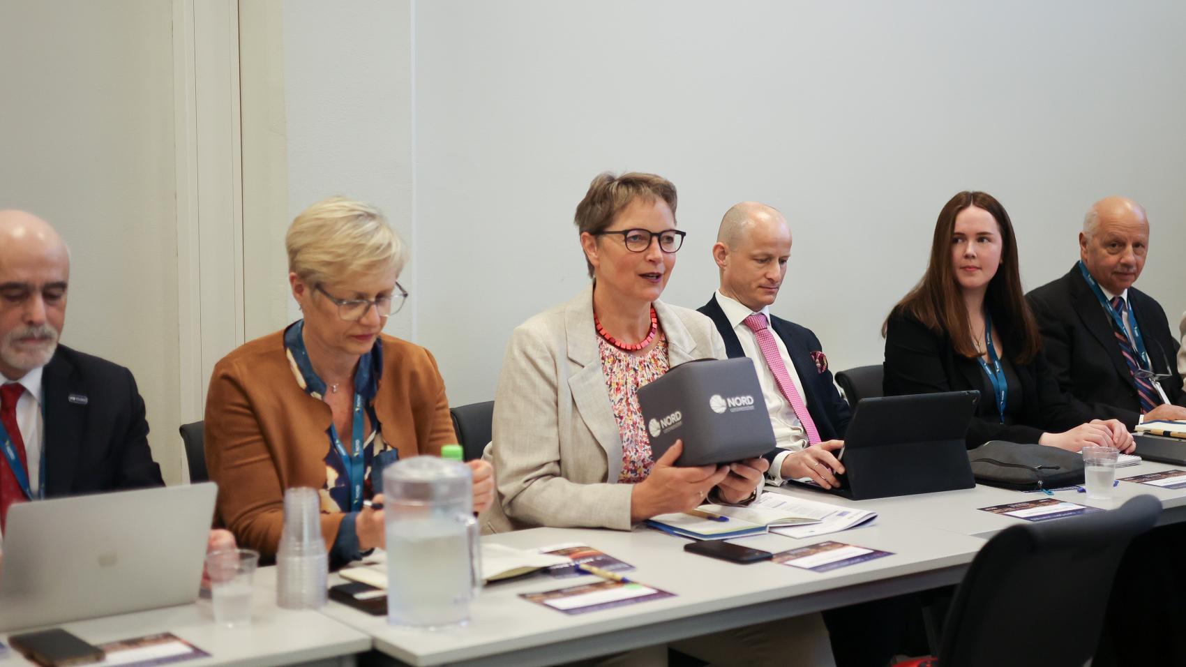 People in a meeting, sitting behind a row of desks. A woman in the middle is talking