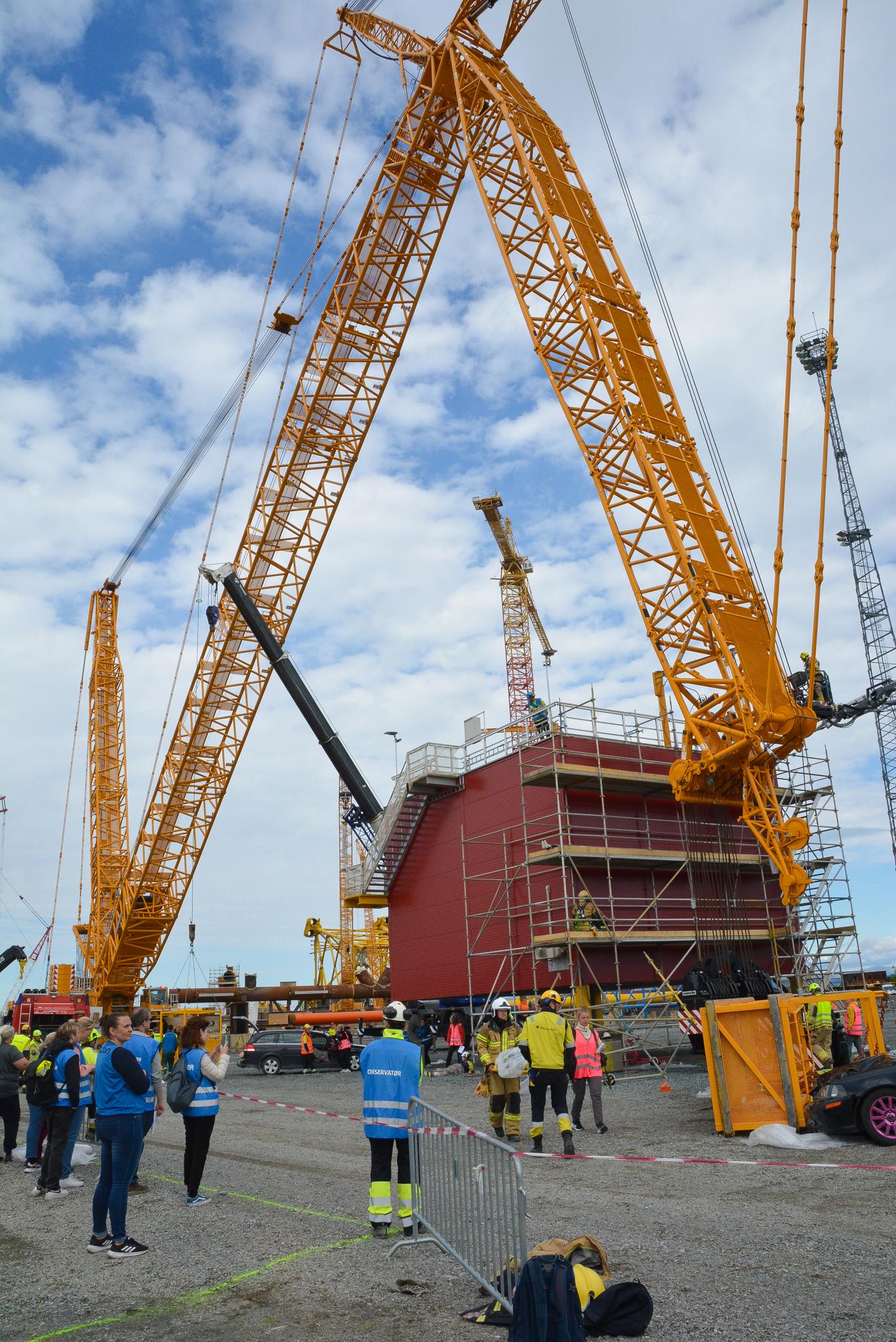 En stor gul kran ligger veltet på et industriområde. Mennesker i blå og gule vester står på området. Foto