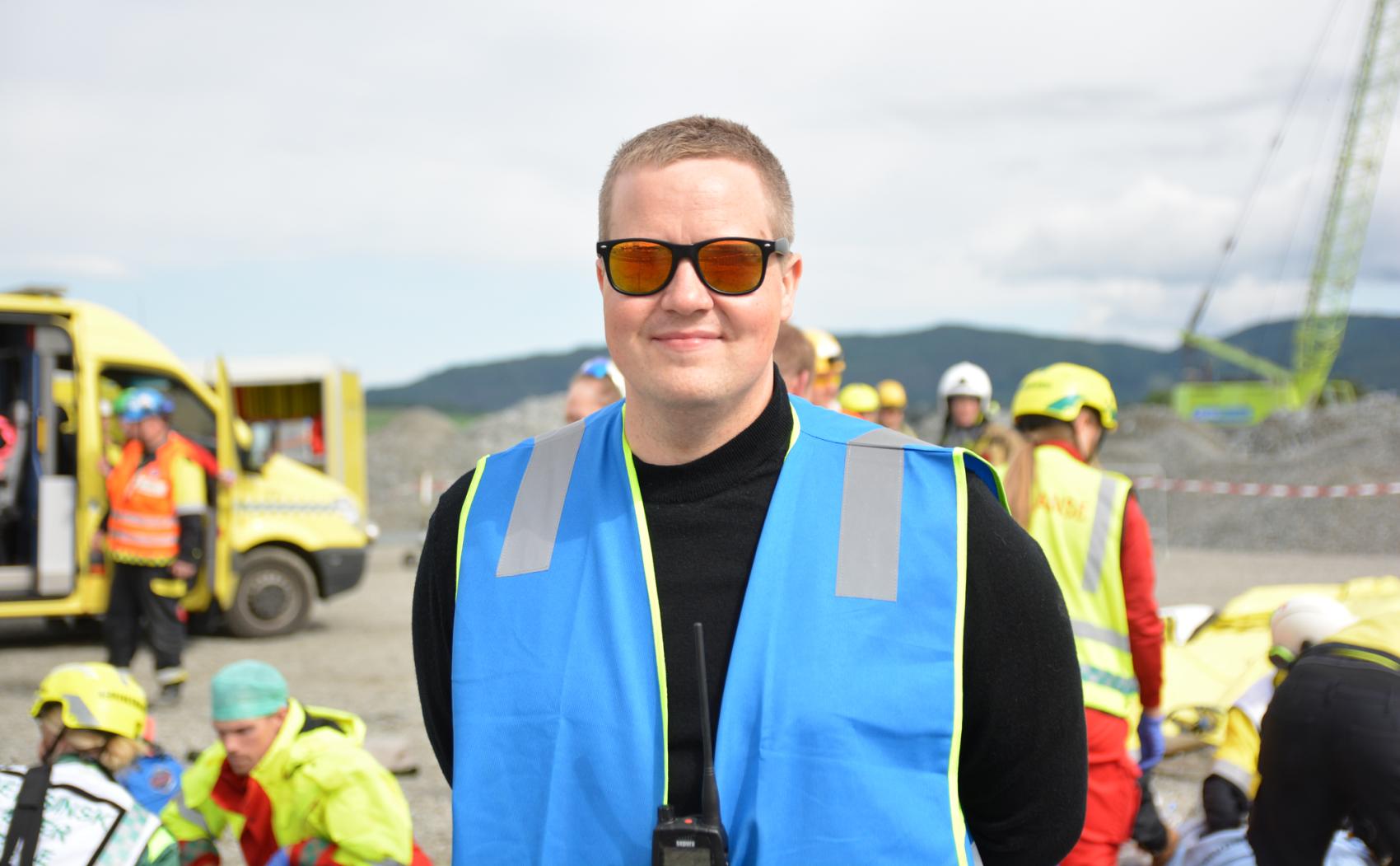Mann med solbruller og blå refleksvest. I bakgrunnen ambulanser og ambulansearbeidere