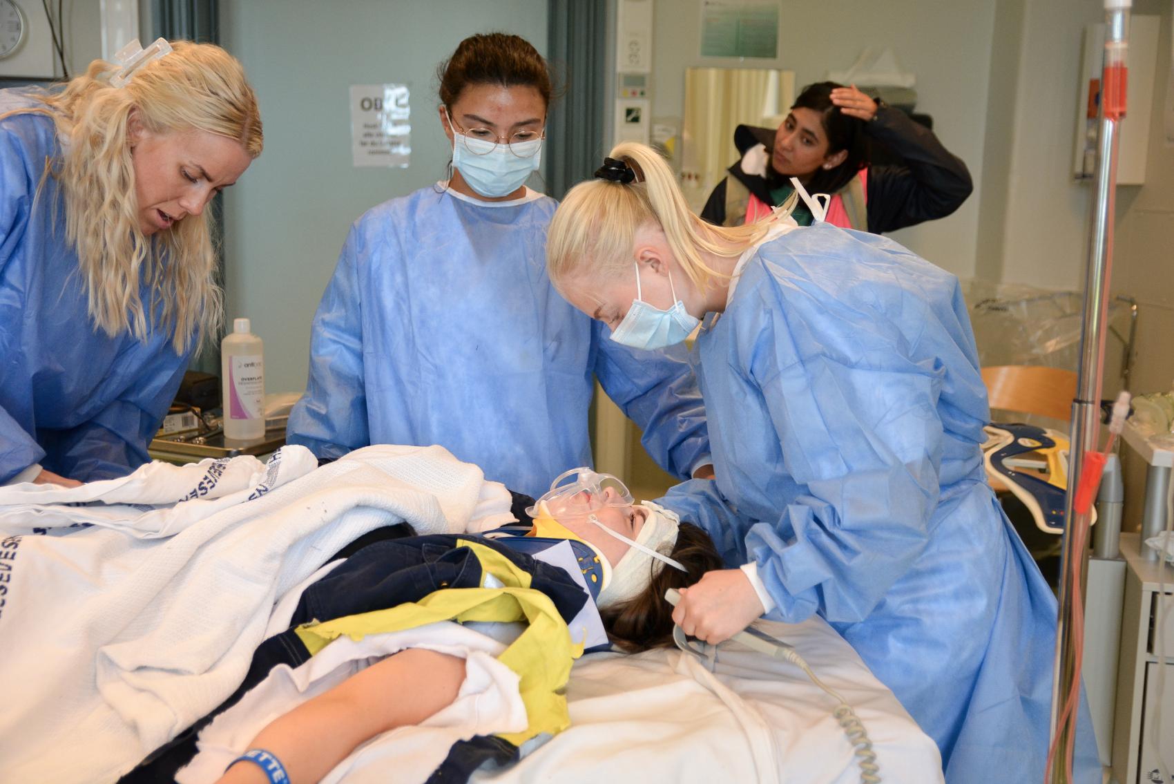 Medisinsk personell i blå drakter og munnbind står over en pasient som ligger i en sykeseng