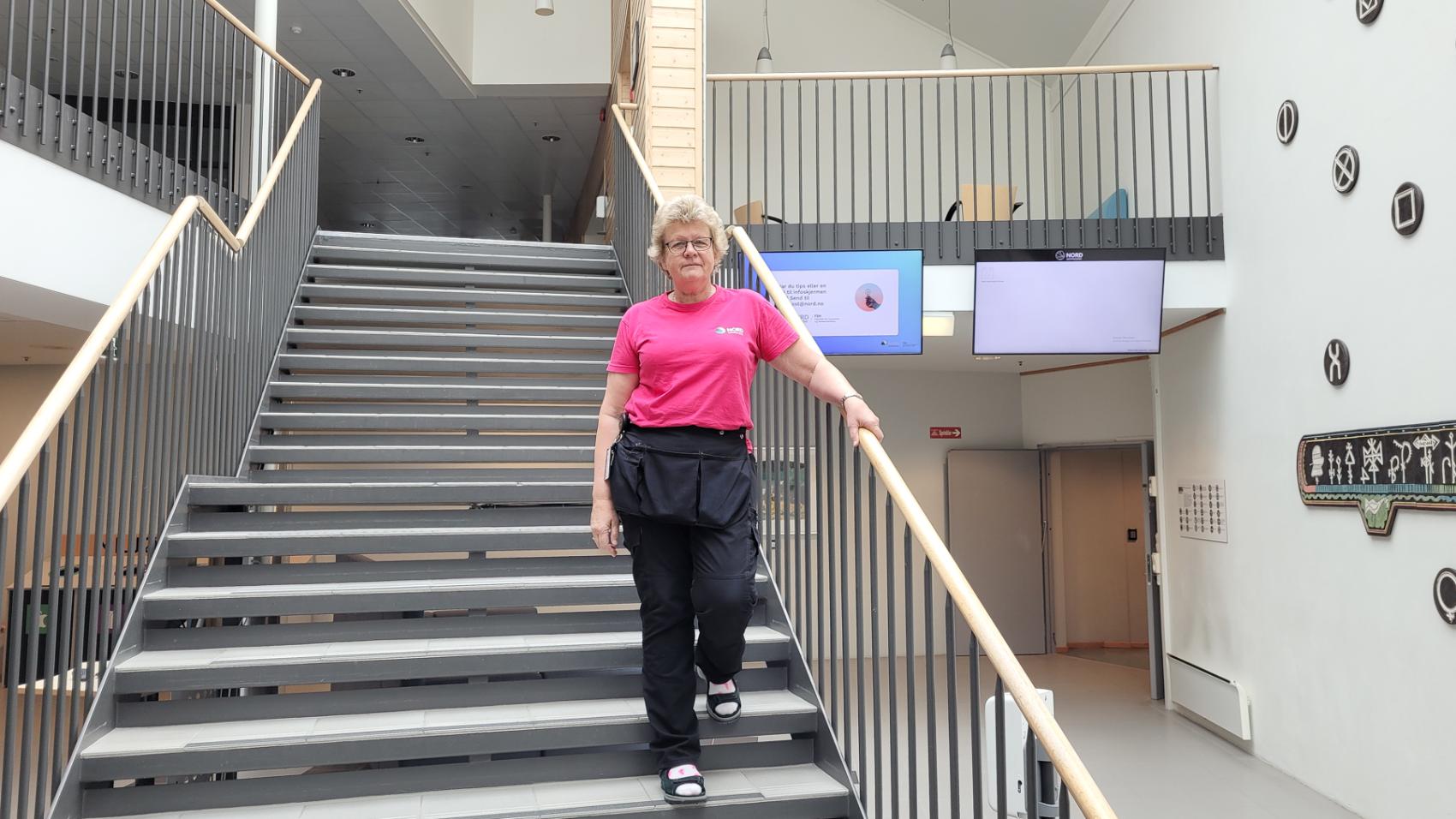 Middelaldrende kvinne i rosa T-skjorte, sort bukse med arbeidslommer, står i en trapp på vei ned i et stort lyst lokale