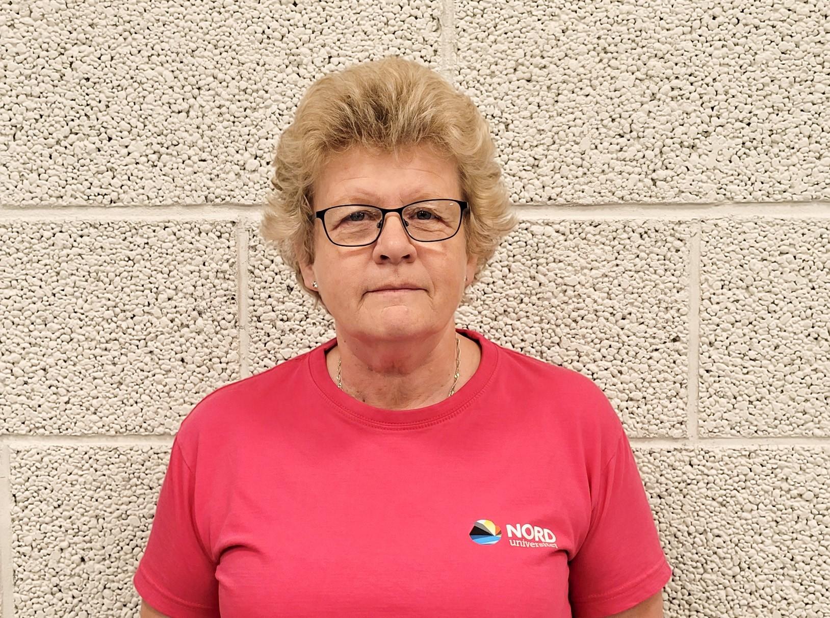Portrettbilde av kvinne med briller, lyst kort hår, kledd i rosa Nord-T-skjorte