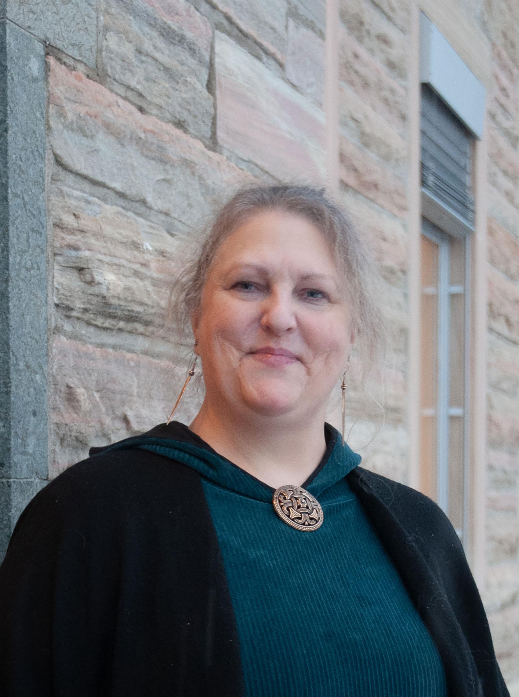 Nærbilde av voksen kvinne foran murbygning. Foto.