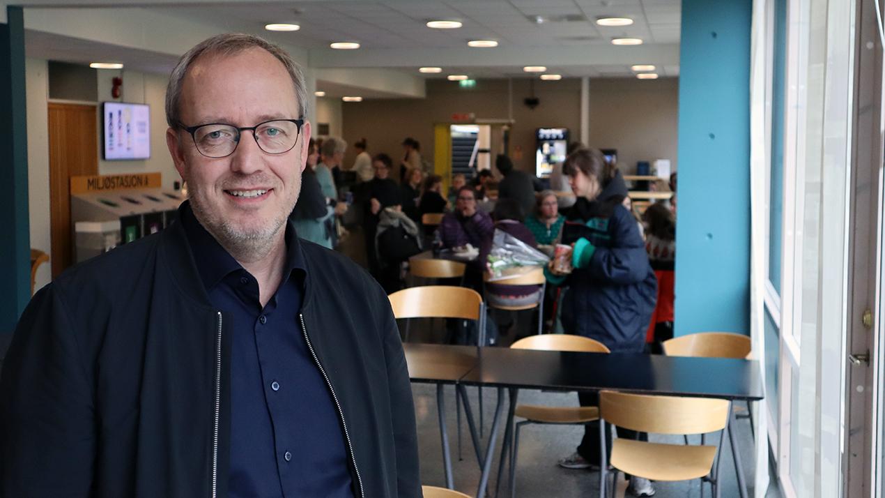 Voksen mann i kantineområde med mennesker i bakgrunn. Veiledningspedagogikk. Foto