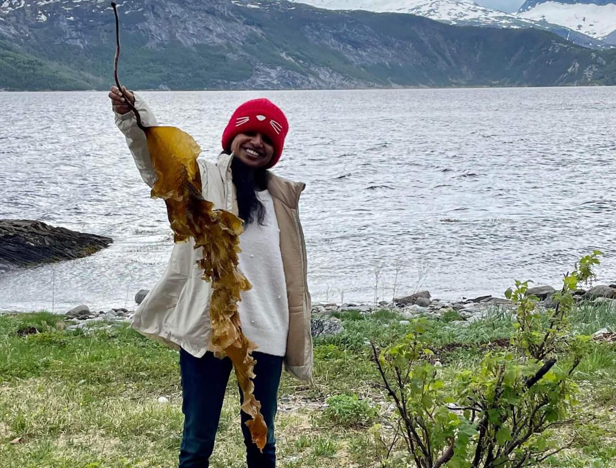 Kvinne med rød lue holder opp tare på strand