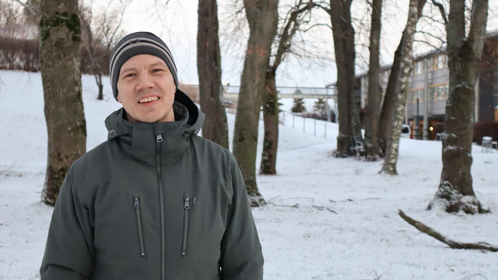 Mann med lue og jakke utendørs, i bakgrunnen trær og grå bygning. Vinter
