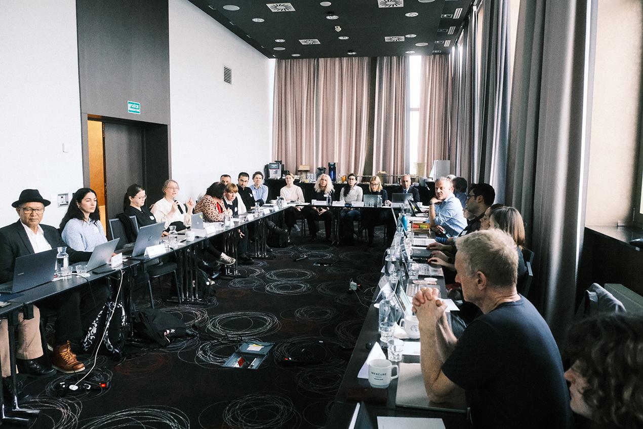 Mange mennesker som sitter rundt et møtebord i en stor møtesal. Foto