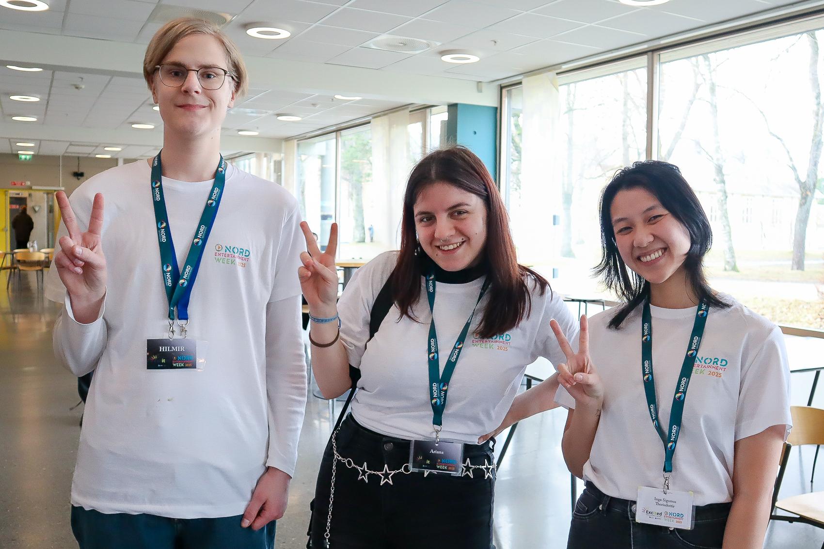 Tre studenter, en gutt og to jenter, i hvite T-skjorter og nøkkelbånd, gjør V-tegn med fingrene.