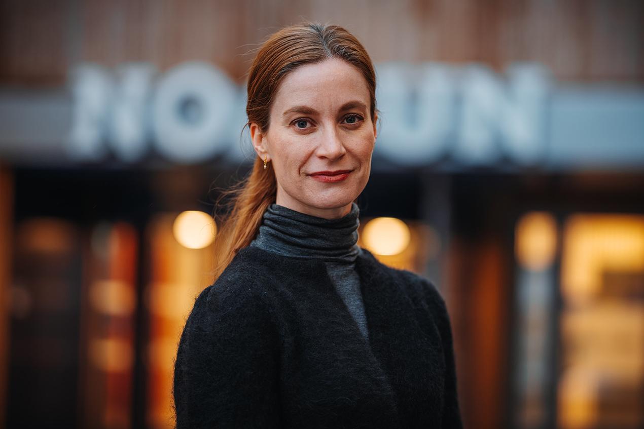 Portrett av en kvinne i sort jakke og høyhalsa genser foran et opplyst bygg. Foto