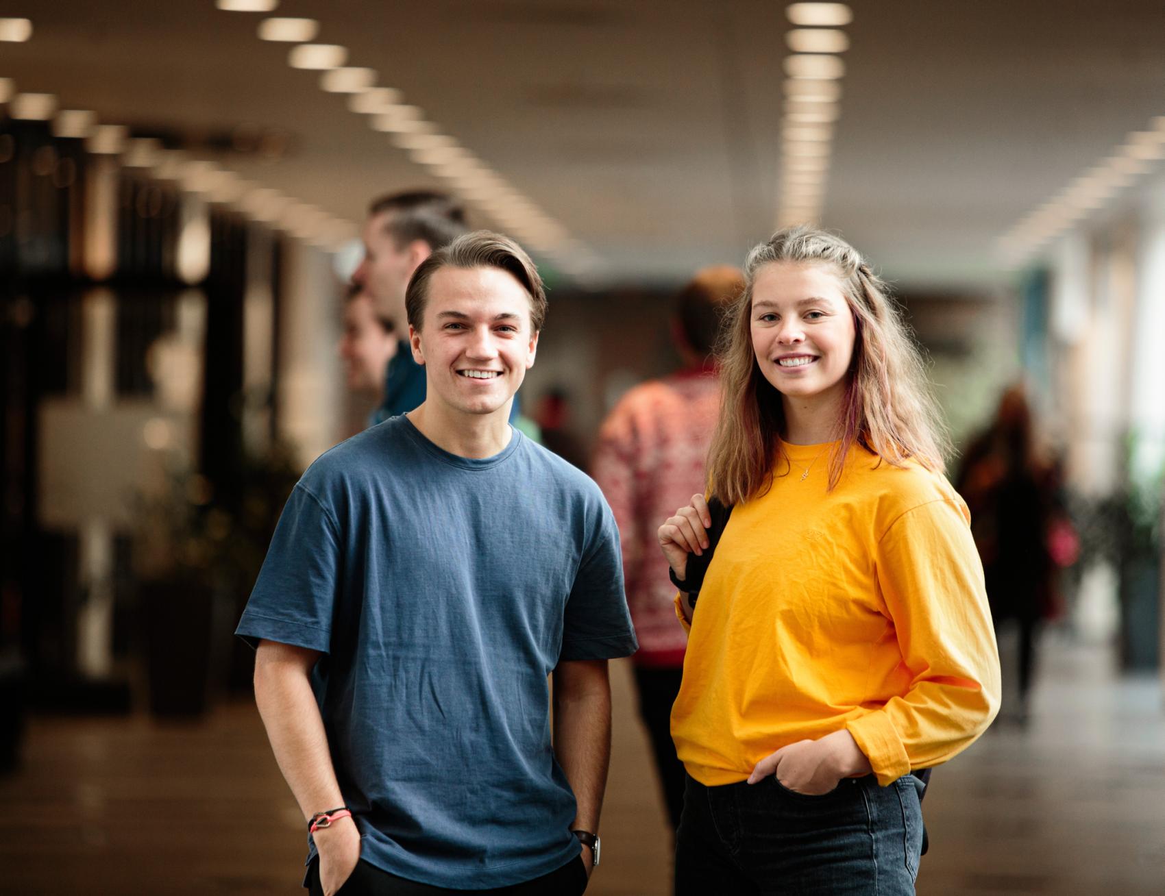 To studenter står i en gang, smiler og ser mot kamera. Foto.