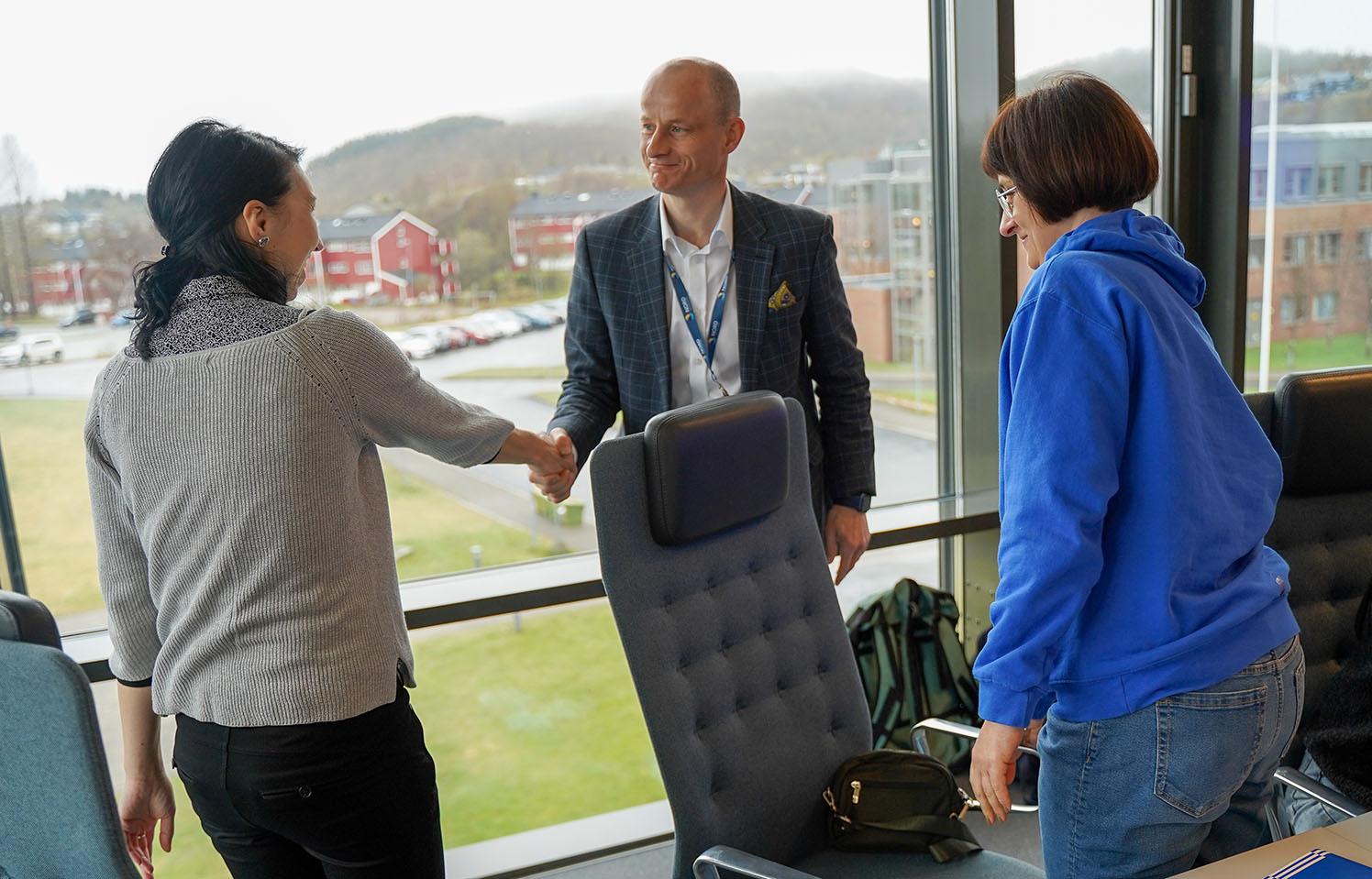 Håndtrykk mellom mann og kvinne, medan ei anna kvinne ser på.