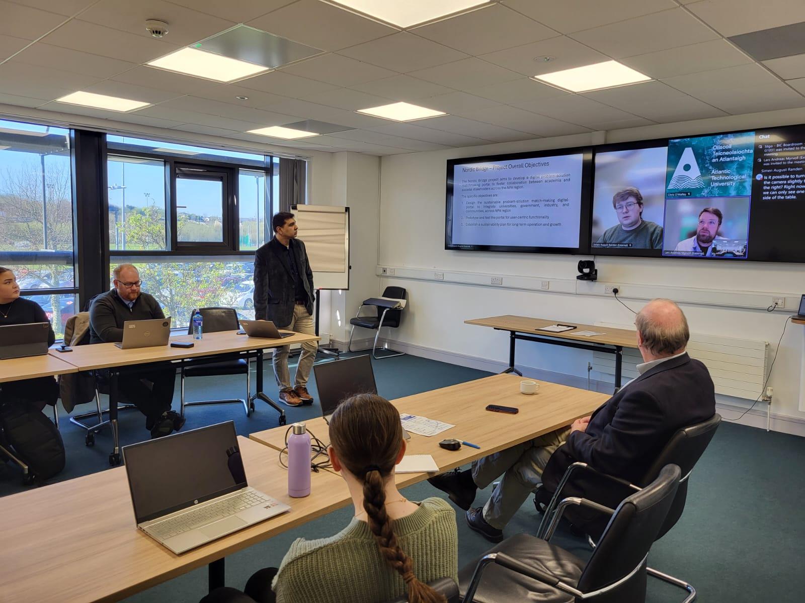 Fem mennesker i et møterom, der møteleder ser på to store skjermer, hvor flere deltar digitalt i møtet.  