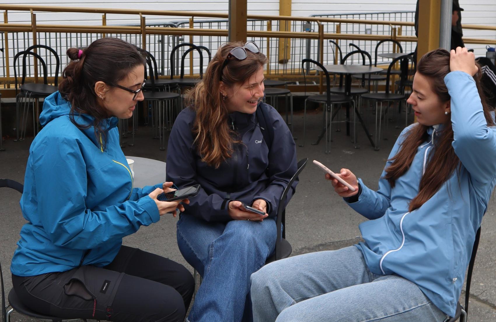 Tre unge kvinner kledd i blått sitter ute og snakker sammen mens de undersøker noe på mobiltelefoner