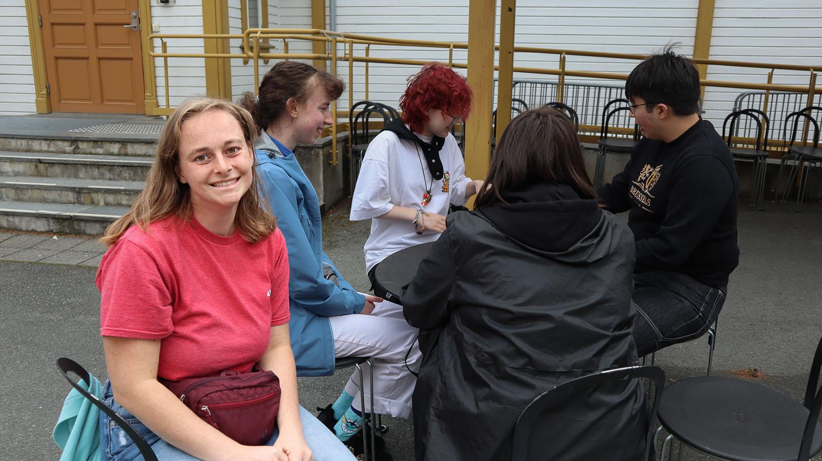 Smilende ung med halvlangt lyst hår og rosa skjorte, sitter på en stol ute, i bakgrunnen fire andre unge som sitter i en ring og snakker sammen