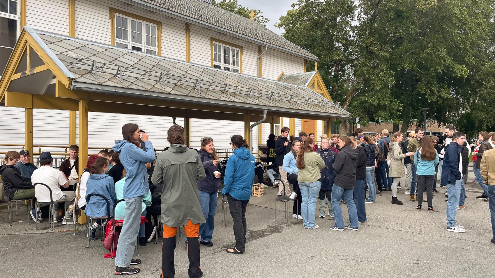 Mange unge mennesker står og sitter utenfor hvit trebygning