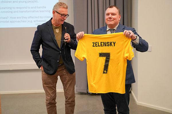 To voksne menn i dressjakket, der den ene holder opp en gul fotballtrøye. Glimtdrakt til Ukrainas president