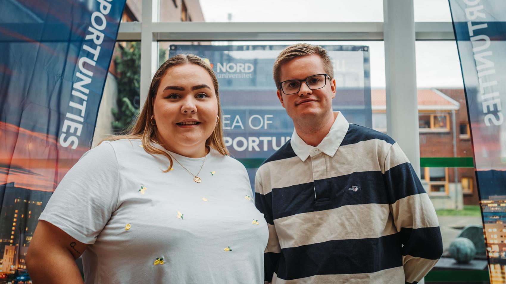 To unge studenter, en mannlig og en kvinnelig, inne foran stor glassfasade. Hanna Mary Pettersen og Hallvard Nøstdahl