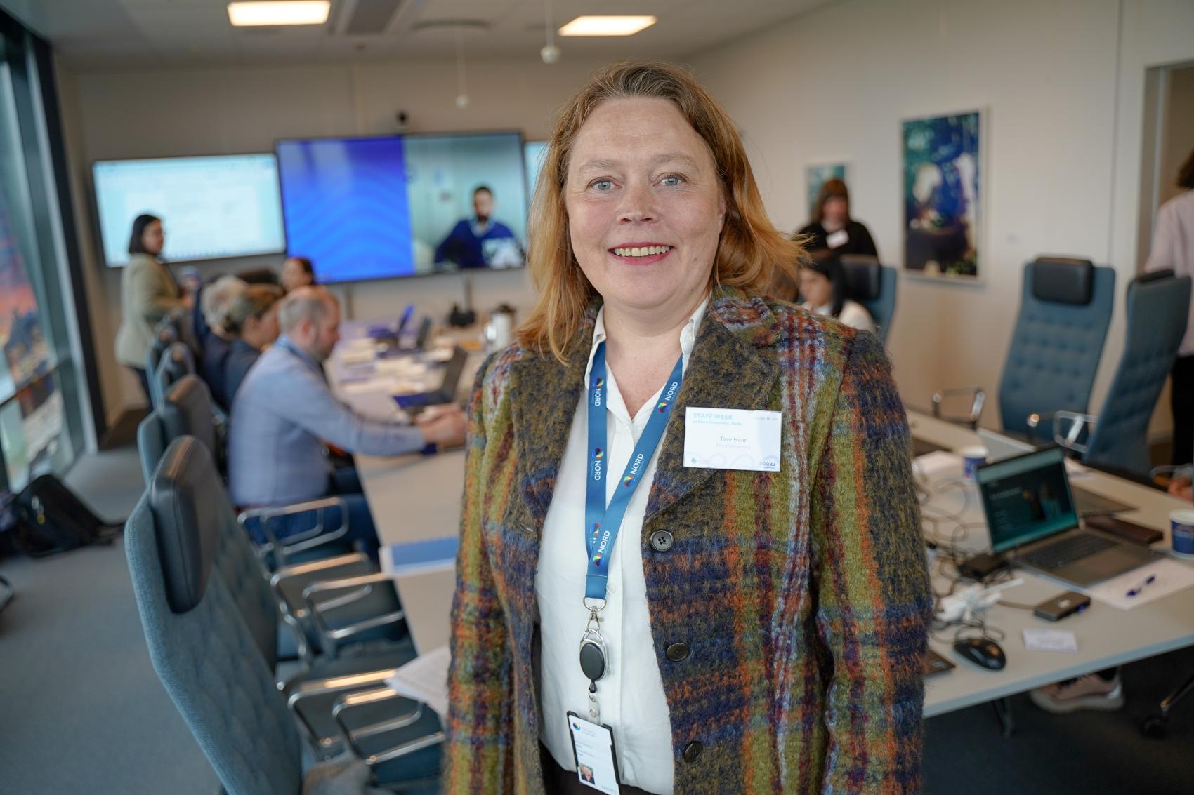 Tove Holm, nærbilde av voksen kvinne i fargerik jakke og med nøkkelkort rundt halsen, inne på et møterom med mange mennesker i bakgrunn.