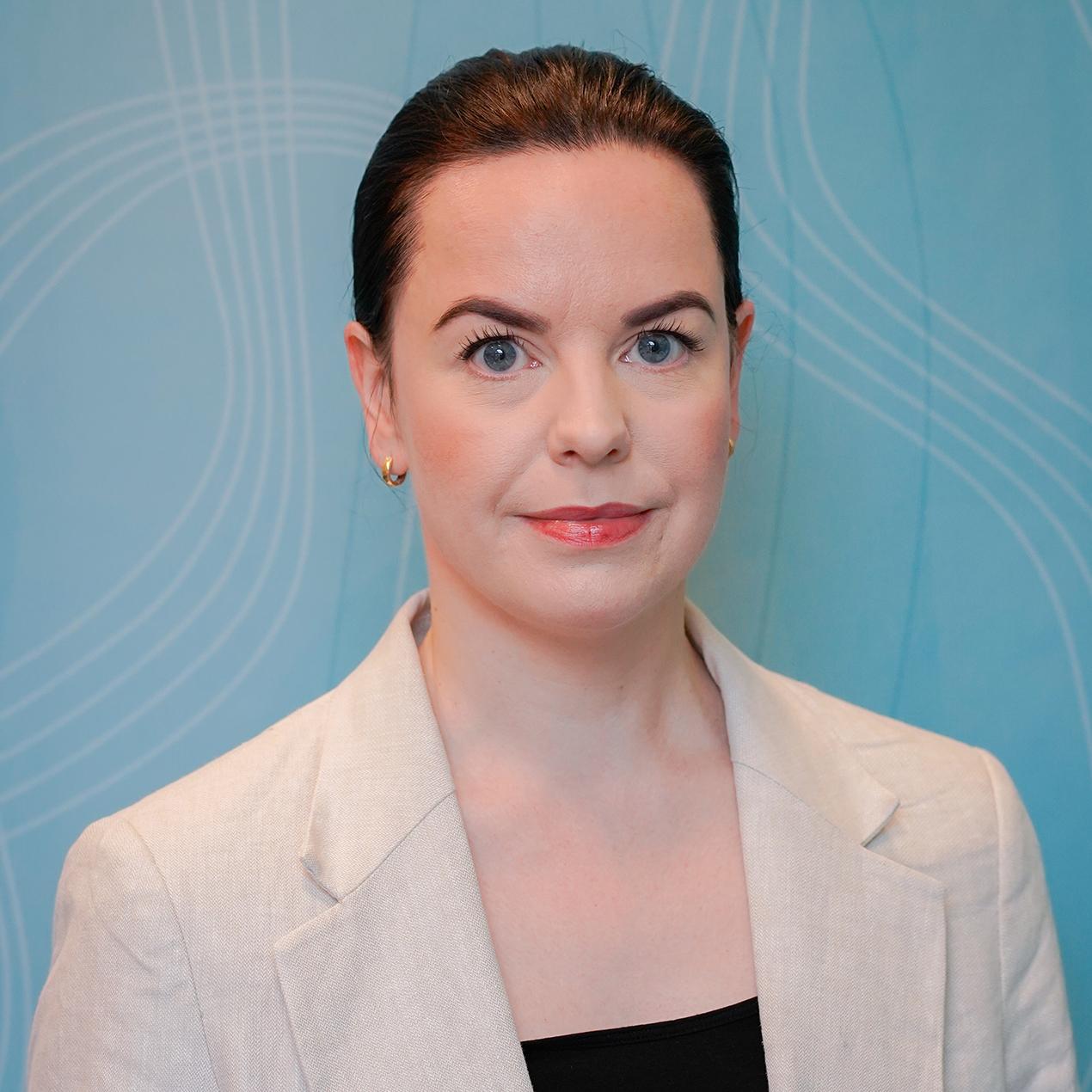 Kvinne med stramt hår og i lys jakke foran lyseblå vegg. Foto